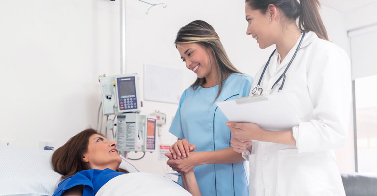 A imagem mostra uma paciente de cabelos castanhos, deitada em uma cama, uma mulher de cabelos claros segurando a mão da paciente e uma médica de jaleco branco, com uma prancheta na mão.