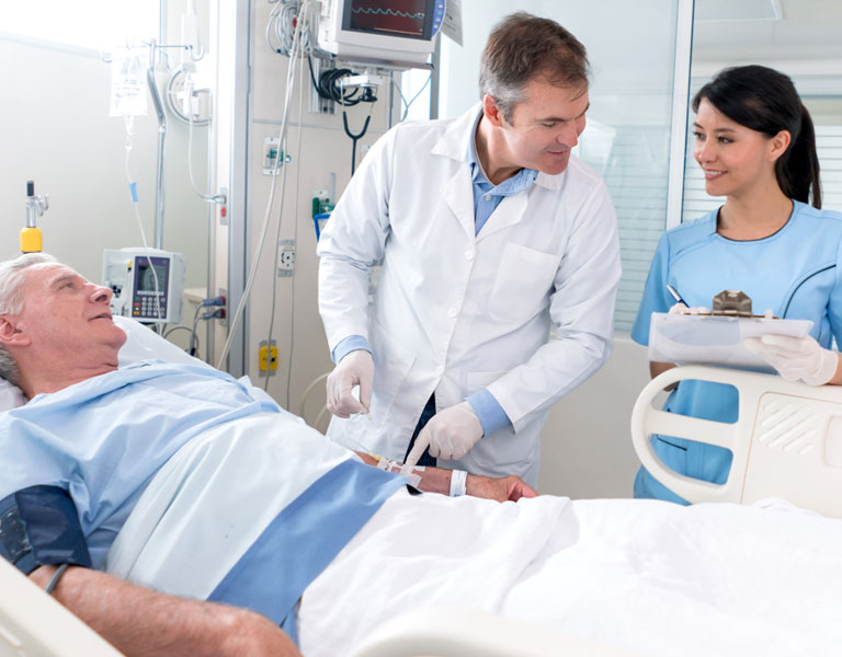 Médico, enfermeira e paciente idoso na sala de internação. O paciente está tranquilo, deitado na maca. O médico olha com leveza para o prontuário que a enfermeira está preenchendo, enquanto conversa com o paciente.