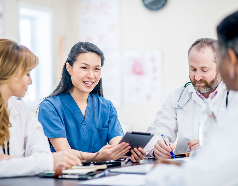 Enfermeira com traços asiáticos e cabelos pretos e lisos fala sorridente com médicos, enquanto segura um prontuário.