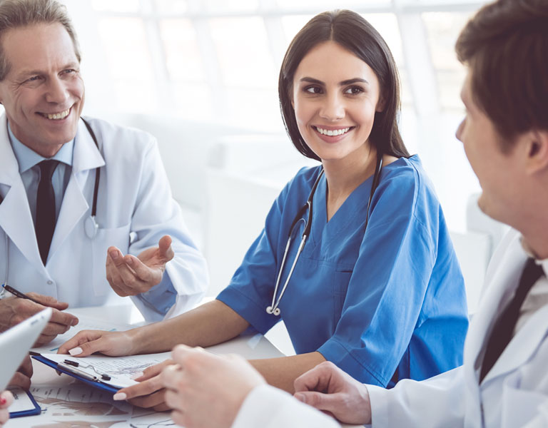 2 médicos de jalecos brancos e uma enfermeira de roupa azul e cabelos castanhos conversam entre si, sorridentes.