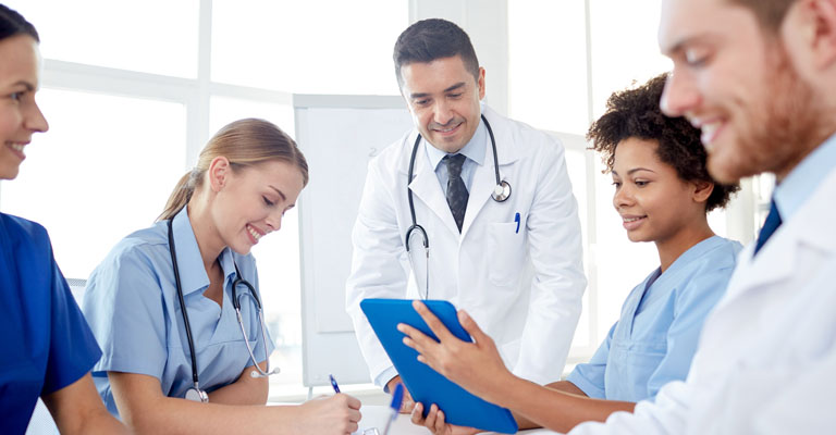 A imagem mostra quatro médicos: duas mulheres e dois homens. Os quatro profissionais estão reunidos e sorriem enquanto observam algo em comum.