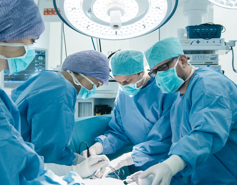A imagem mostra um centro cirúrgico com duas médicas e dois médicos, todos com máscara, touca, luvas e aventais cirúrgicos, realizando um procedimento ao redor de uma maca.